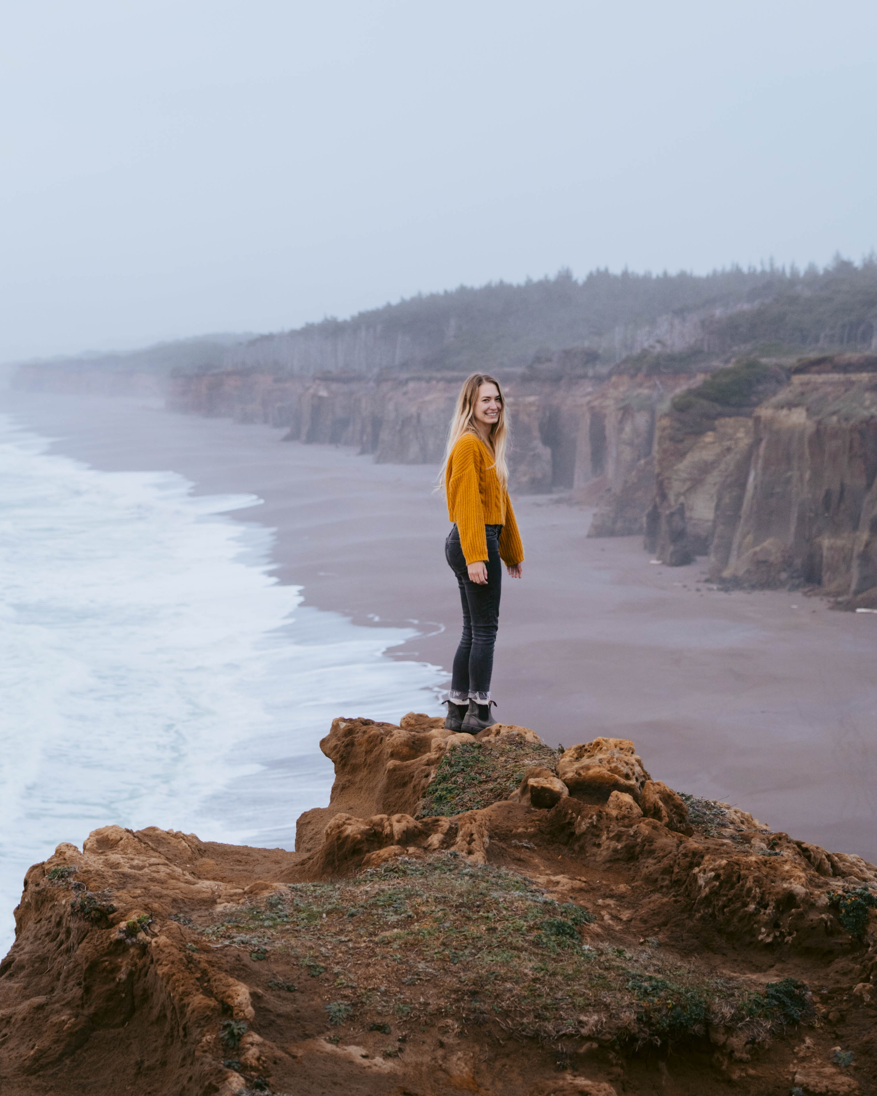 Oregon coast hikes near Oregon Treehouse. A woman stands on a cliff ledge that overlooks the vast Oregon coast with cliffs on the side. You can see the waves creating patterns miles down the coast. 